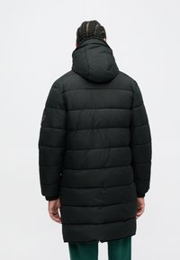 Άτομο με πλεξούδες που φοράει μακριά μαύρη φουσκωτή ζακέτα με κουκούλα, στέκεται στραμμένο μακριά, μπροστά σε απλό φωτεινό φόντο.