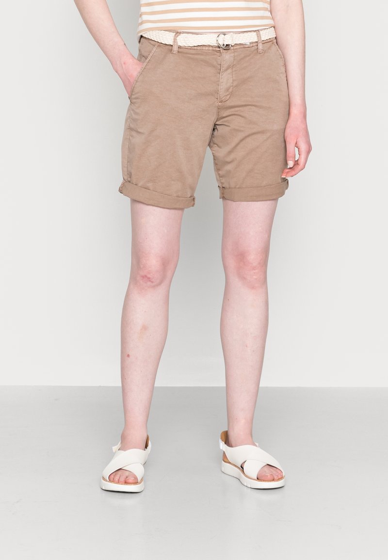 Personne portant un short marron clair retroussé avec une ceinture tressée, des sandales blanches à lanières croisées et une chemise beige rayée, debout sur un fond uni.