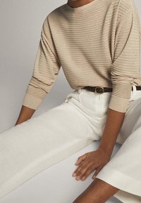Maglione beige a coste con collo rotondo, abbinato a pantaloni bianchi a vita alta. Visibile una cintura nera e dettagli in tessuto testurizzato.