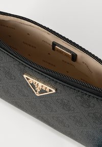 Czarny teksturowany portfel na zamek z złotym trójkątnym logo. Wnętrze w kolorze beżowym z powtarzającym się wzorem "GUESS". Gładkie zamknięcie na zamek.