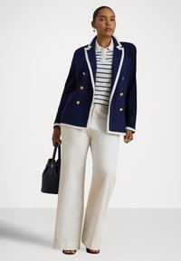 Moteris, apsirengusi tamsiai mėlynu švarku su balta apdaila, dėvi dryžuotą baltą polo marškinėlį, platėjusias kreminės spalvos kelnes, laiko juodą rankinę, stovi priešais paprastą foną.