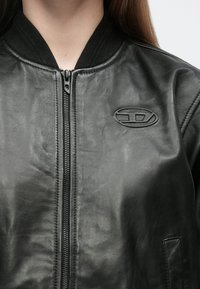 Czarna skórzana kurtka bomberka z zamkiem z przodu, czarnym prążkowanym kołnierzem i teksturowaną naszywką z logo na lewej piersi. Gładkie wykończenie.