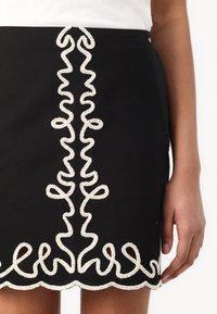 Jupe mini noire avec broderie blanche à volants le long de l'avant et de l'ourlet. Présente une texture lisse et une silhouette ajustée et épurée.
