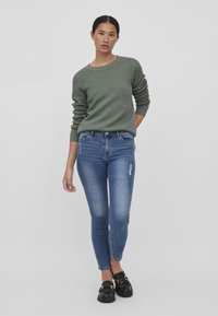 Pull en maille verte, jeans bleus ajustés avec légères éraflures, mocassins noirs avec des accents en or, se tenant sur un fond neutre.
