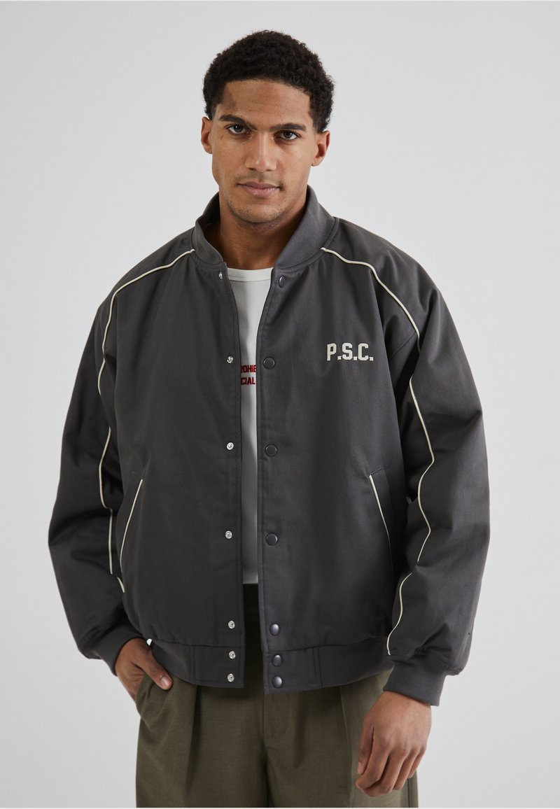 Jachetă bomber gri cu dungi albe, închidere cu butoane tip snap și logo-ul P.S.C. pe partea stângă a pieptului. Textură moale, design minimalist.