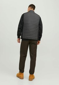 Grau gesteppte Weste mit hohem Kragen, getragen über einem schwarzen Sweatshirt. Kombiniert mit braunen Hosen und tanfarbigen Stiefeln, von hinten gesehen.