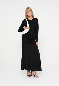 Schwarz langärmliges Oberteil mit ausgestelltem Saum, kombiniert mit einem schwarzen gerafften Rock. Das Model hält eine weiße Handtasche und trägt schwarze Riemchen-Heels.