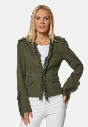 Olivgrüne Jacke mit Fransenverzierung am Kragen und an den Ärmeln, zwei Fronttaschen und taillierter Passform. Getragen über einem weißen Oberteil.