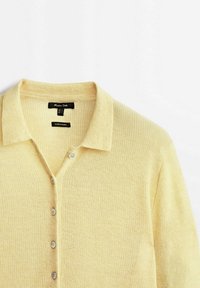 Cardigan en tricot jaune clair avec col et quatre boutons, présenté à plat sur un fond blanc.