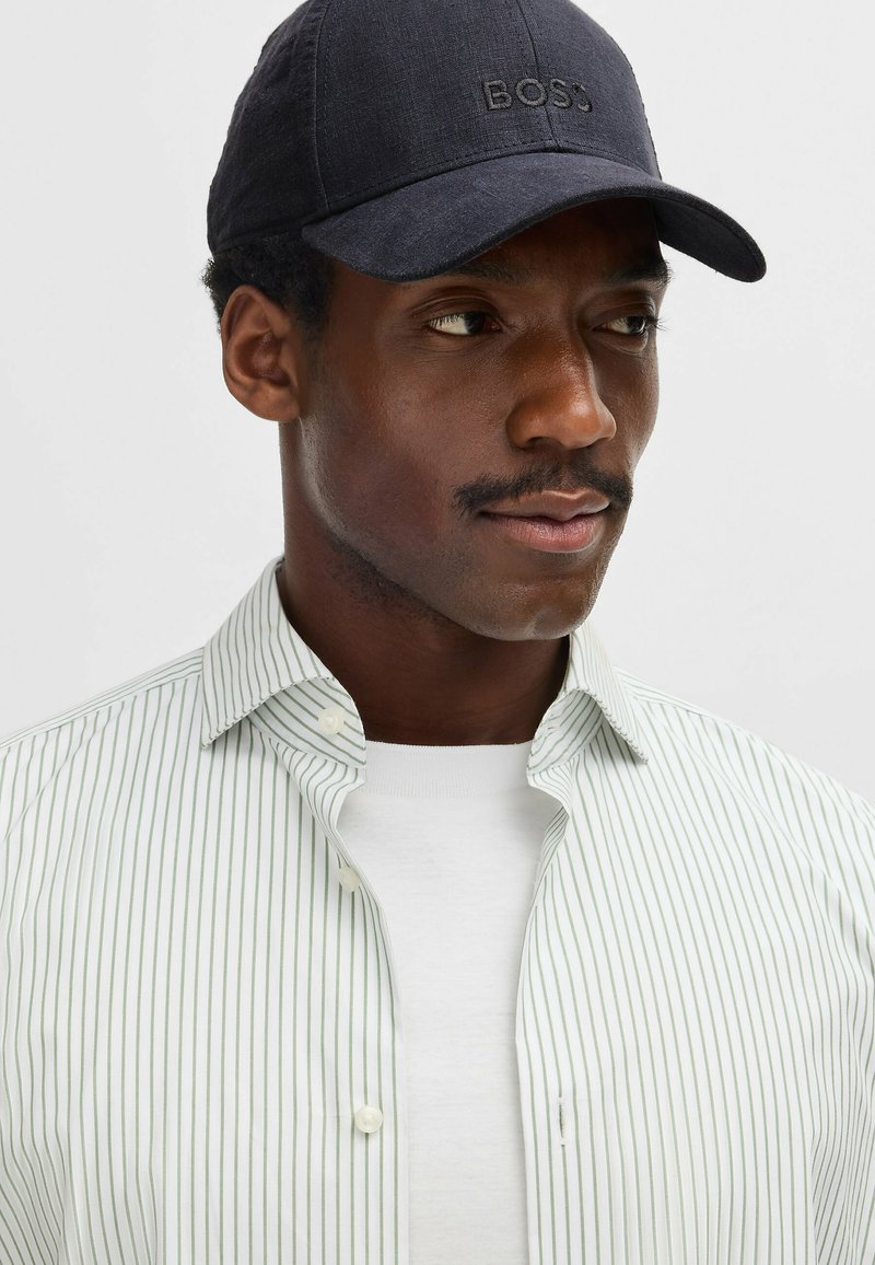 Navy cap with "BOSS" logo, green and white striped button-up shirt, white crew neck t-shirt underneath. Smooth fabric and tailored fit.