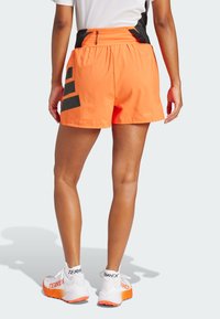 Pantalones cortos deportivos naranjas con un diseño de acento negro, que cuentan con rayas en los paneles laterales. La persona está usando zapatillas blancas y naranjas.