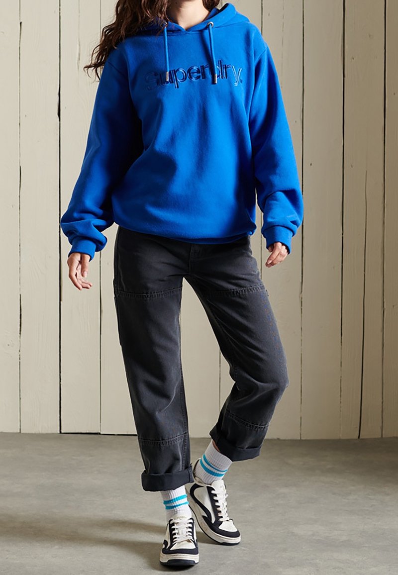 Personne portant un sweat à capuche Superdry bleu vif, un pantalon noir retroussé, des chaussettes blanches à rayures bleues et des baskets noir et blanc, debout à l'intérieur.