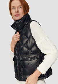 Schwarze, gesteppte Steppweste mit hohem Kragen, glänzender Oberfläche und Vordertasche. Trägt man über einem strukturierten weißen Pullover mit gerippten Bündchen.