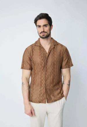 Chemise à manches courtes marron à motifs avec une texture ondulée et un col, dotée de boutons sur le devant, portée avec un pantalon de couleur claire.