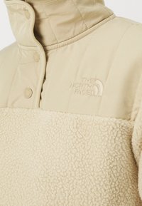 Beżowy polarowy sweter z pikowaną górą z poliestru, wysoki kołnierz, zapięcie na zatrzaski oraz haftowane logo na piersi. Miękka tekstura.