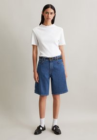 Weißes, geripptes T-Shirt mit gewelltem Saum, kombiniert mit blauen Denim-Shorts und einem schwarzen Gürtel. Schwarze Loafers und weiße Knöchelsocken runden den Look ab.