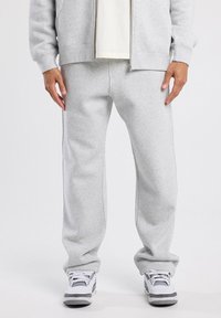 Cuerpo inferior de una persona que lleva pantalones de chándal gris claro, chaqueta con cremallera a juego, zapatillas blancas con detalles en gris y negro, de pie sobre un fondo blanco.