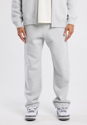 Ala ala bawah seseorang yang mengenakan celana sweat abu-abu muda, jaket zip serasi, sepatu sneakers putih dengan detail abu-abu dan hitam, berdiri di latar belakang putih.
