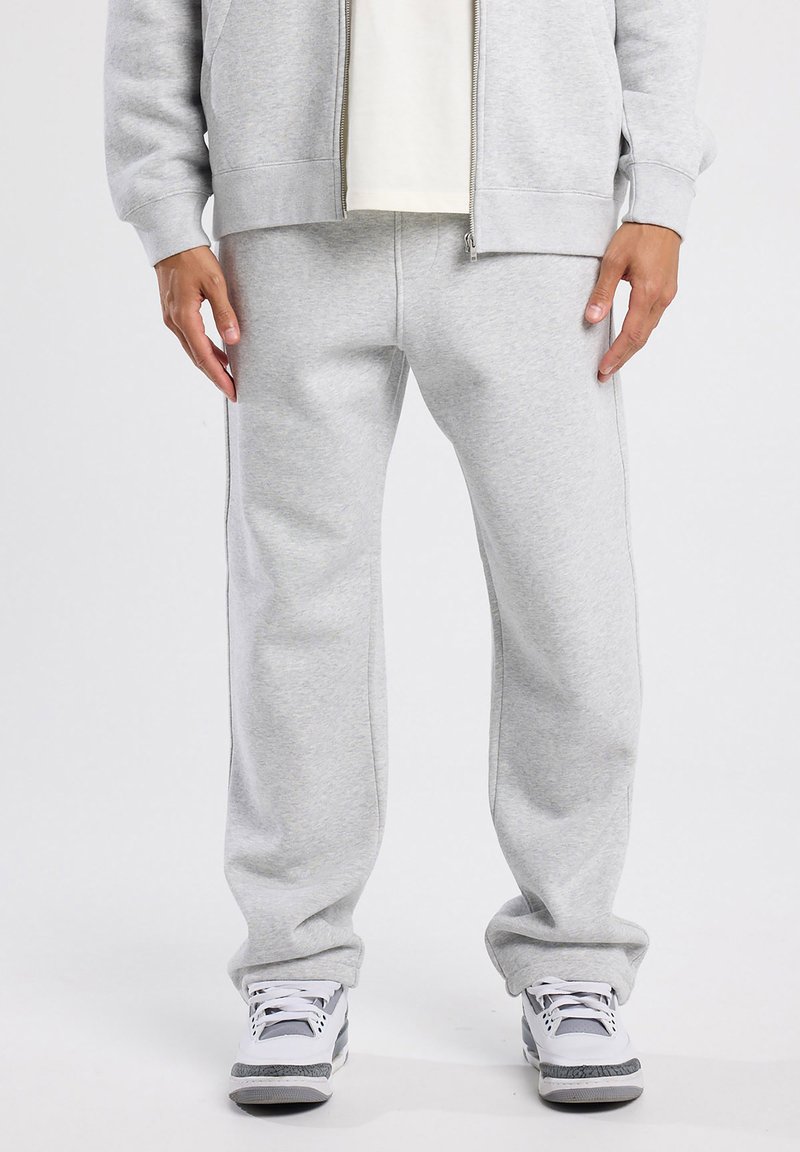 Cuerpo inferior de una persona que lleva pantalones de chándal gris claro, chaqueta con cremallera a juego, zapatillas blancas con detalles en gris y negro, de pie sobre un fondo blanco.