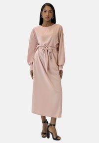 Lange, zachte roze jurk met een losse pasvorm, getailleerd met een ceintuur in de taille. Voorzien van lange mouwen en een eenvoudige ronde halslijn.