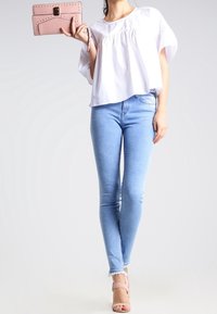 Blusa bianca a maniche corte con dettagli arricciati, jeans skinny azzurri chiari e pochette rosa pallido. La modella indossa sandali nudi con cinturini.