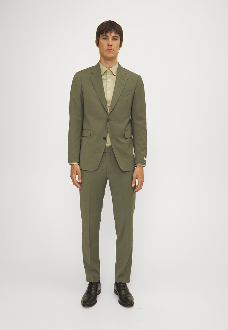 Olivgrüner Anzug mit slim fit, einreihiger Blazer und geraden Hosen, kombiniert mit einem hellbeigen Hemd und schwarzen Schuhen.