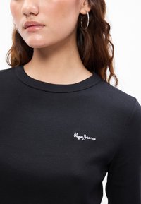 Fekete bordás, hosszú ujjú póló, kerek nyakkivágással és fehér hímzett logóval a mellkasán. A modellen ezüst karika fülbevalók vannak.