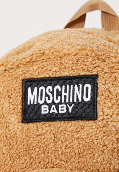 Sac à dos en tissu marron fluffy avec une étiquette rectangulaire noire affichant "MOSCHINO BABY" en texte blanc. Comprend une boucle de suspension en haut.
