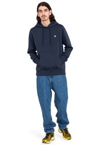 Sudadera azul oscura con bolsillo frontal, cordones ajustables y pequeño logotipo en el pecho. Combinada con unos vaqueros holgados de color azul claro y zapatillas amarillas.