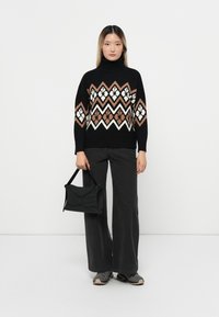 Schwarzer Pullover mit einem Zickzackmuster in Braun und Creme, hoher Rollkragen, kombiniert mit grauen Weitbeinen und einer schwarzen Handtasche.