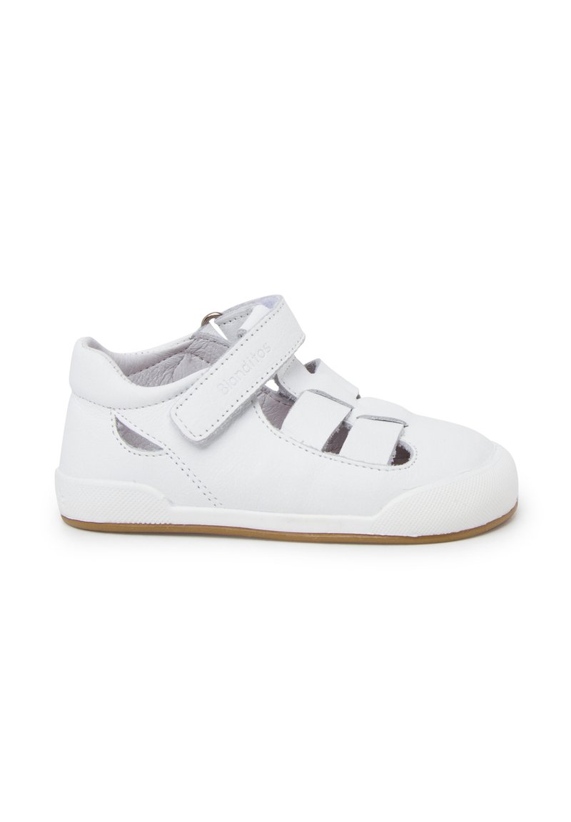 Weißer Lederschuh mit glatter Textur, geschlossener Zehenpartie, verstellbarem Klettverschluss und durchbrochenen Seiten. Gummisohle mit hellbraunen Akzenten.