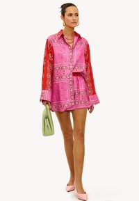 Vestido camisero de manga larga colorido en patrones de paisley rosa y rojo, ajustado en la cintura, combinado con un bolso verde claro y zapatos rosas.