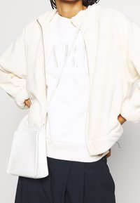 Bílá fleecová bunda na zip s oversized střih, žebrovanými manžetami, doplněná bílým svetrem s reliéfními písmeny a malou bílou kabelkou.