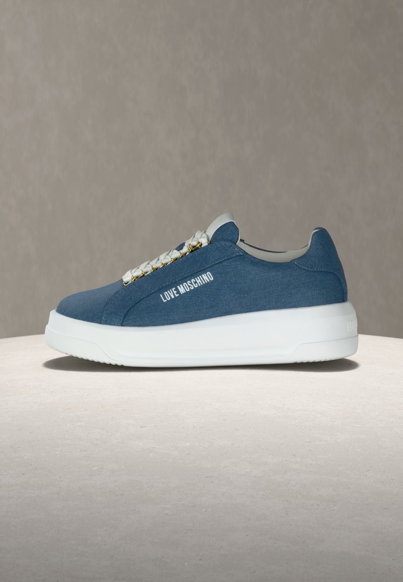 Baskets Love Moschino en denim bleu avec semelle blanche épaisse et lacets blancs à accents dorés, présentées sur une surface en pierre claire.