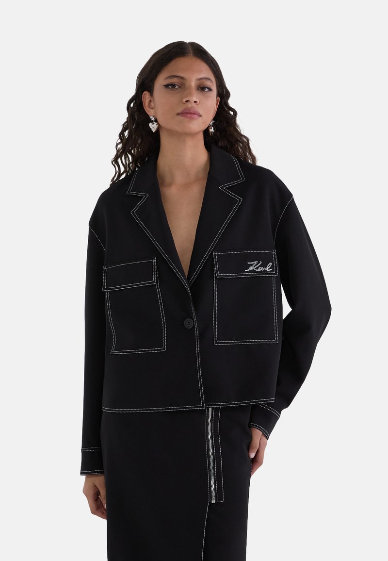 Zwarte cropped blazer met witte stiksels, twee voorzakken, een enkele knoopsluiting en een geknipt kraag. Gladde stoftextuur.
