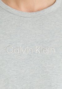 Grijze katoenen T-shirt met korte mouwen en een ronde hals. Bevat witte geborduurde tekst "Calvin Klein" op de borst.