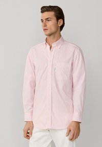 Chemise à manches longues à rayures horizontales rose clair et blanc, avec col à boutons et poche avant. Tissu en coton, coupe décontractée.