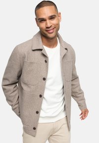 Veste en mélange de laine brun avec col, fermeture à boutons et deux poches poitrine. Stylée avec une chemise blanche en dessous et un pantalon clair.