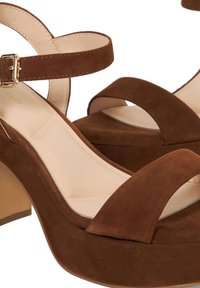 Talons compensés en daim marron avec un talon carré et une sangle à la cheville, présentant un bout ouvert arrondi et une doublure intérieure lisse. Détails en métal doré.