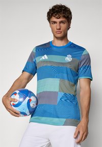 Blauw grafisch voetbalshirt met verschillende textuurpatronen; speler houdt een blauw en witte voetbal vast met rode accenten.