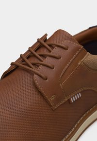 Chaussure habillée en cuir marron avec un motif texturé, lacets marron, détails cousus et accents en tissu beige près du talon.