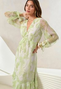 Rochie transparentă, până la podea, în verde pal, cu modele florale, margine volanată și mâneci voluminoase. Prezintă un decolteu adânc în formă de V.