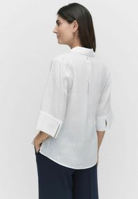 Hvid linned skjorte med 3/4 ærmer, krave, knapdetalje bagpå og slidser i siderne. Stoffet har et blødt, tekstureret udseende.
