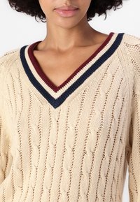 Nainen käyttää kermaista päällikudos-neuletta, jossa on laivastonsininen, kerma ja burgundinpunainen raidoitettu kaulus, joka paljastaa ylärinnan ja kaulan.