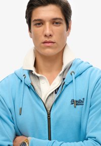 Sudadera con cremallera de color azul claro y cremallera dorada, sobre una camisa gris con cuello. Presenta un logo bordado en el pecho.