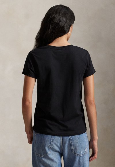 Γυναίκα με μακριά σκούρα μαλλιά, που φοράει ένα απλό μαύρο κοντομάνικο μπλουζάκι και ανοιχτό μπλε τζιν, στέκεται με την πλάτη σε ένα μονόχρωμο φόντο.