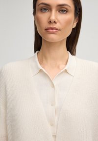 Donna con lunghi capelli scuri che indossa una camicia crema a righe con bottoni e un cardigan cremoso testurizzato, su uno sfondo chiaro e semplice.