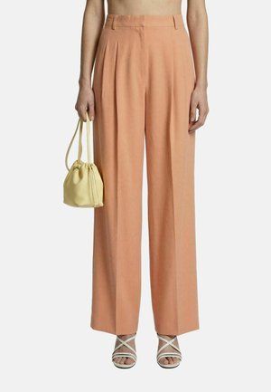 Donna che indossa pantaloni color pesca a vita alta e gamba larga, tiene una piccola borsa gialla, indossa sandali bianchi con cinturini, inquadrata fino al torso.