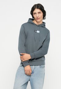 Felpa con cappuccio di colore grigio ardesia, caratterizzata da un patch con logo frontale, polsini a coste e una vestibilità rilassata. Abbinate a jeans azzurri chiari.