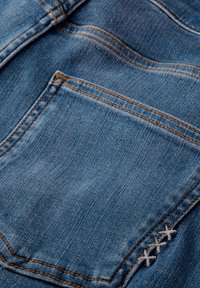 Tejido de mezclilla con un tono azul, que presenta un bolsillo trasero y costuras en contraste, con acentos decorativos de bordado en forma de X blanca.